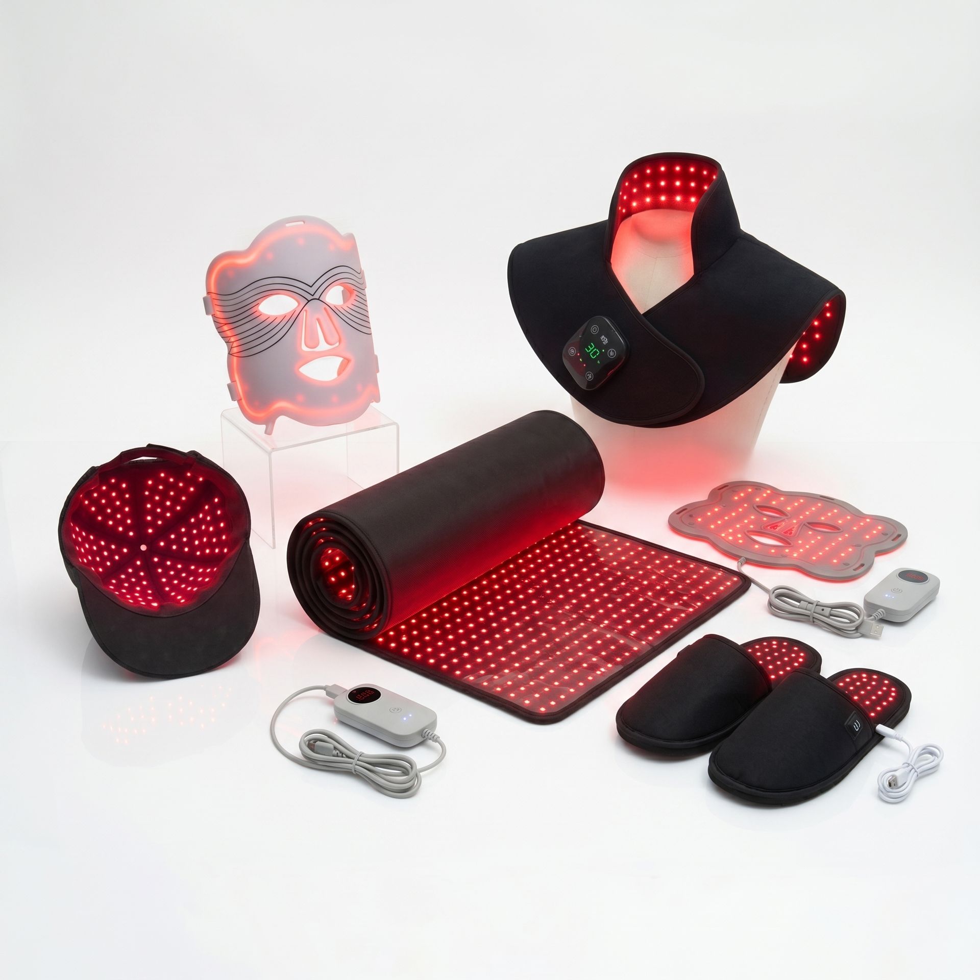 Assorted red-light therapy devices laid out on a white surface, including a neck wrap, a rolled LED mat, illuminated face masks, and control units ready for use